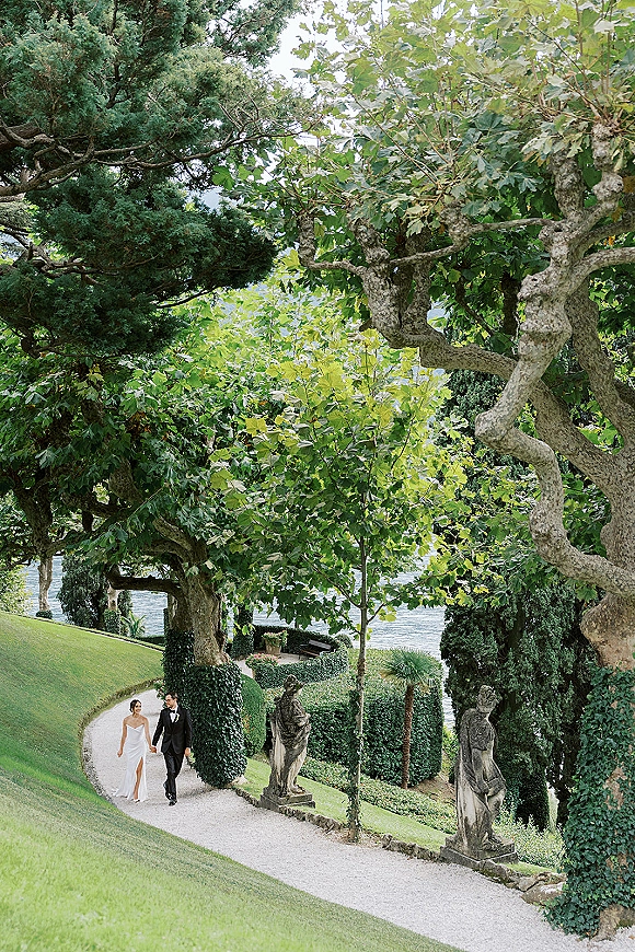 Couple portrait of bride and groom walking hand in hand, her strapless dress and bouquet beside a lakeside garden path with statues