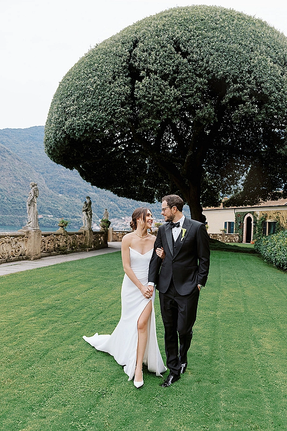 Couple portrait of bride and groom walking hand in hand, her strapless slit gown and pearl necklace on a villa garden terrace by lake
