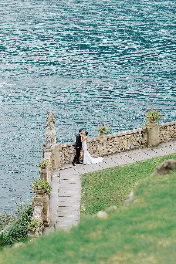 Wedding kiss portrait of bride and groom kissing on a stone terrace with ocean water behind, framed by a statue and potted flowers