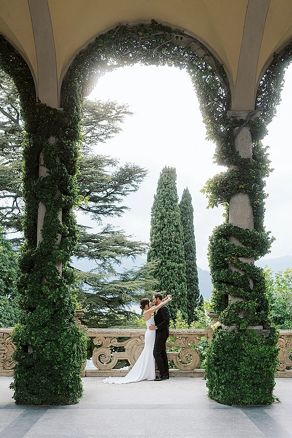 Wedding kiss portrait of bride and groom kiss under an ivy-covered stone arch on a villa terrace with lake and mountain backdrop
