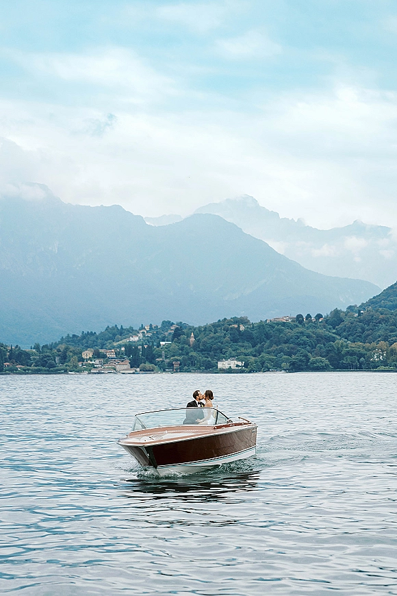 Wedding kiss portrait of newlyweds on boat, bride in veil and groom in tuxedo on a motorboat with mountain lake and village backdrop