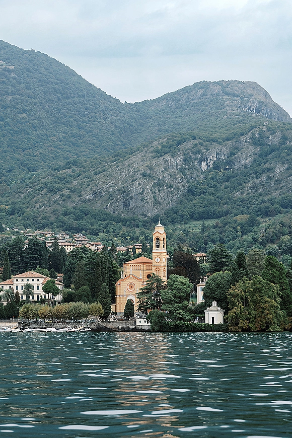 Mountain lake landscape with a yellow lakeside church and clock tower beside shoreline buildings, framed by forested hills and cloudy sky