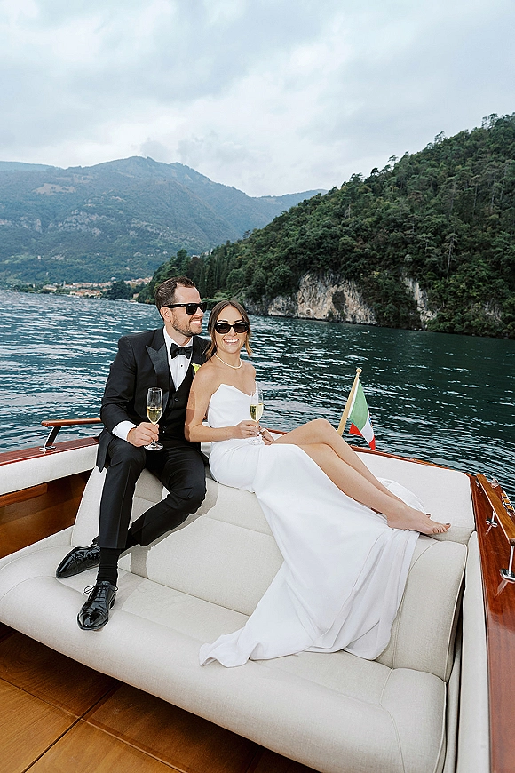 Couple portrait of bride and groom in sunglasses on a boat wedding photos, raising champagne flutes on a lake with mountains behind