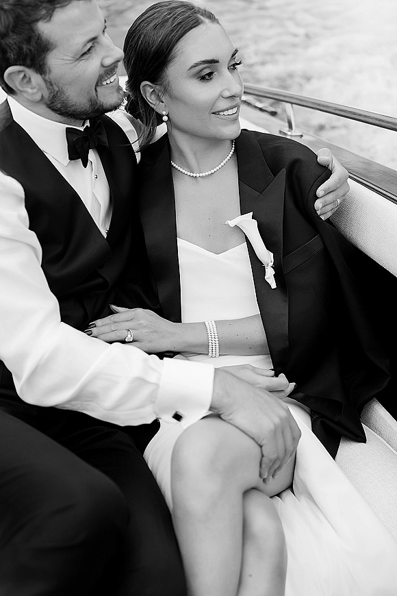 Couple portrait in a black and white wedding portrait style, bride in blazer and pearls leaning on groom in tuxedo on boat by water
