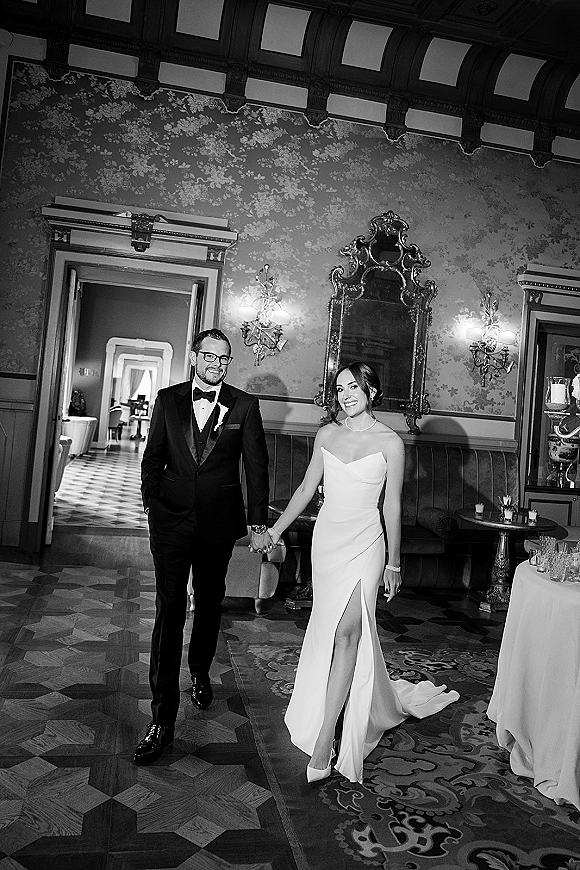 Couple portrait of bride and groom holding hands, her strapless slit gown and his tux with glasses, before ornate mirror and sconces indoors