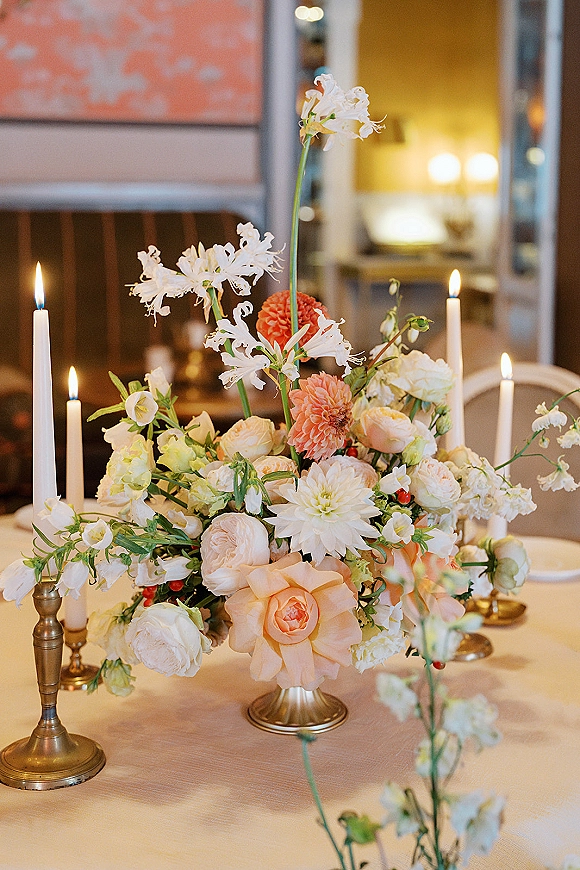 Wedding centerpiece with pastel floral arrangement and brass candlesticks, taper candles glowing on a table in an indoor reception space