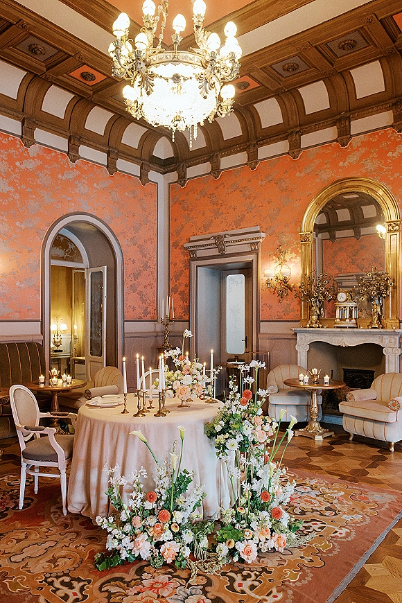 Reception tablescape with round wedding table decor on a blush tablecloth, brass candles and floral centerpieces beneath an ornate chandelier