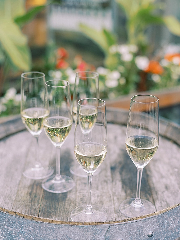 Champagne glasses filled with sparkling wine on a wooden barrel table, surrounded by greenery and flowers at a rustic wedding station