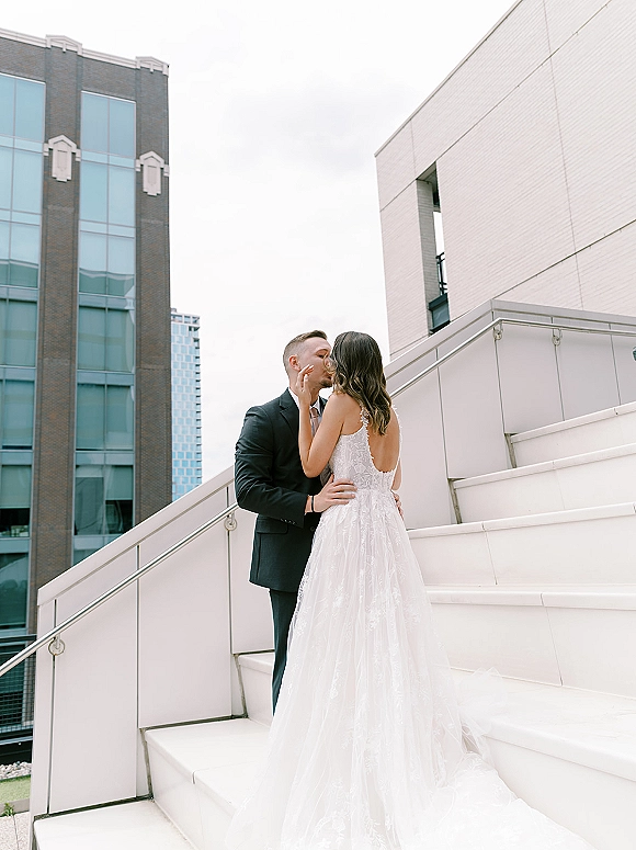 Wedding kiss portrait of bride and groom kissing on outdoor stairs, her open-back lace gown against modern buildings and skyline backdrop
