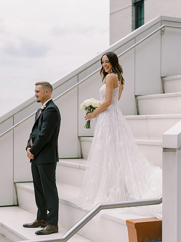Wedding first look on stairs as bride approaches groom, holding a white rose bouquet in a lace dress, modern building behind