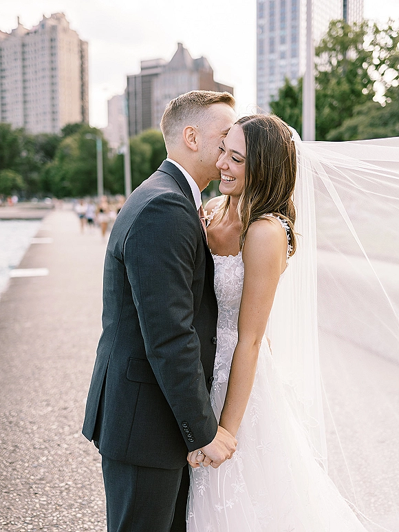 Couple portrait of bride and groom holding hands as he kisses her forehead, veil blowing in the wind along a waterfront skyline walkway