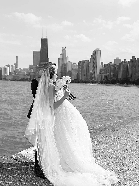 Couple portrait in a black and white wedding portrait style, bride and groom embracing on a concrete pier with city skyline beyond the lake