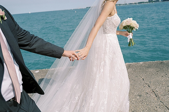 Couple portrait of bride and groom holding hands, her veil blowing as she carries a white rose bouquet on a waterfront pier by the ocean horizon