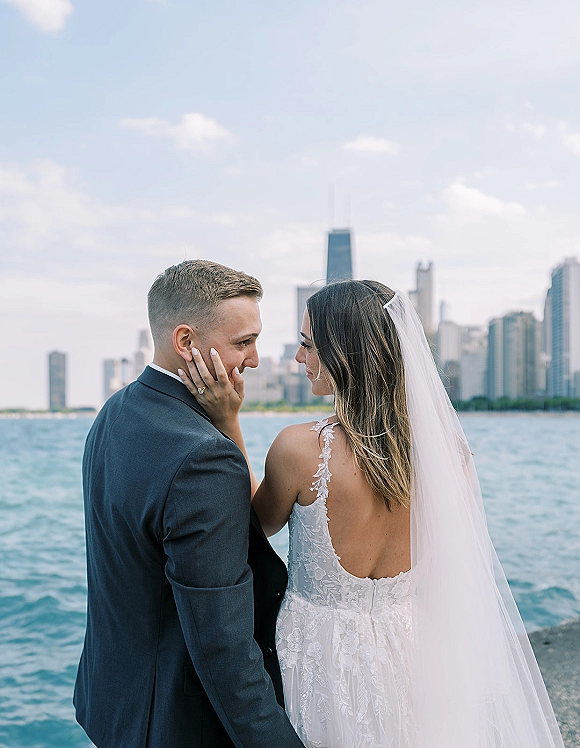 Couple portrait with bride touching groom’s face as they stand by the waterfront, city skyline behind them, her veil flowing in the breeze
