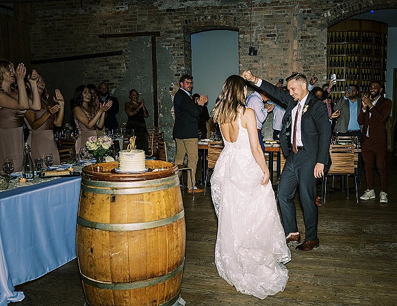 First dance as bride in wedding dress and groom in suit twirl before an exposed brick wall with wine bottle display and guests watching