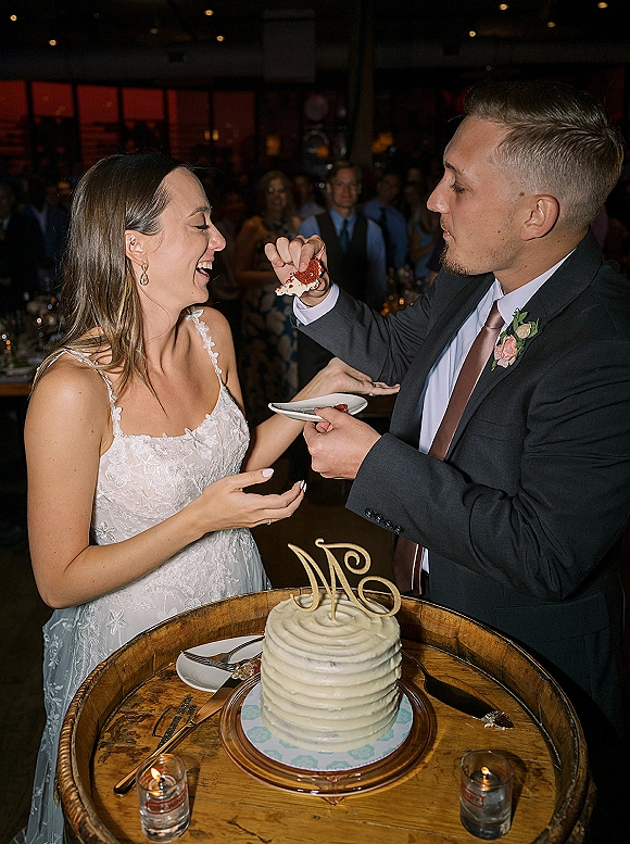 Cake cutting moment as bride and groom feed each other a bite beside a white buttercream wedding cake on a candlelit wooden table under string lights