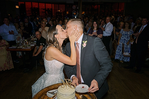 Wedding cake cutting as the bride and groom share a kiss beside a white buttercream cake with initials topper, guests cheering behind