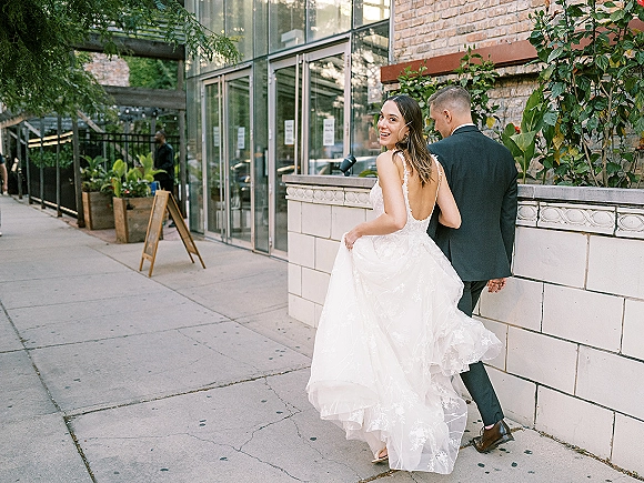 Couple portrait of bride looking back while walking with groom in black suit, lace open-back gown trailing along city sidewalk by brick wall