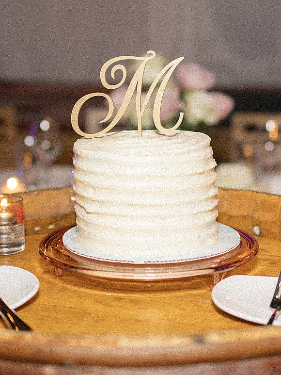 Wedding cake with white buttercream cake frosting on a stand, topped with a monogram M, surrounded by votive candles on a wood table