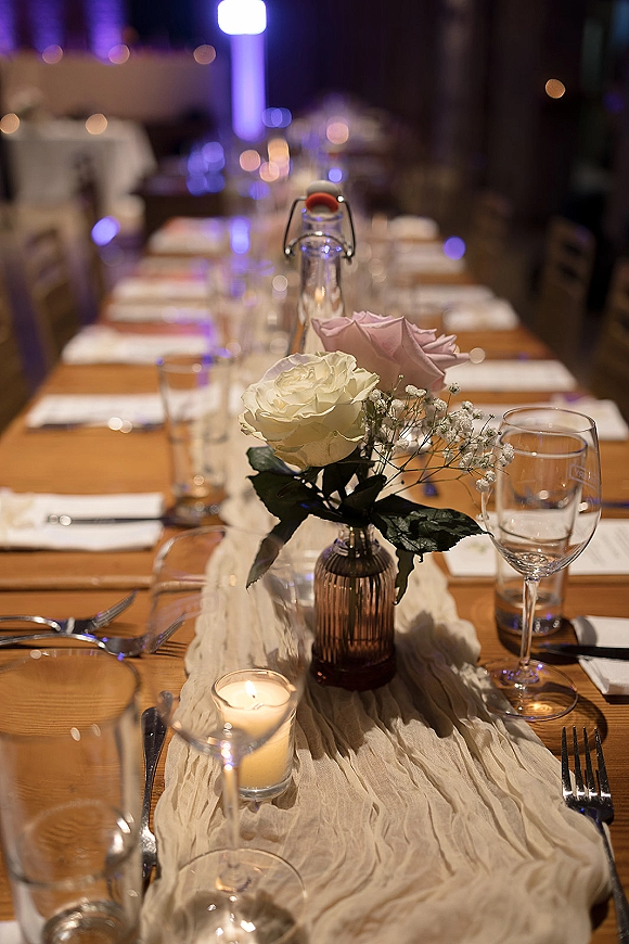 Reception tablescape on a rustic banquet table with gauze runner, amber vase of roses and baby's breath, candles, and glassware in dim light