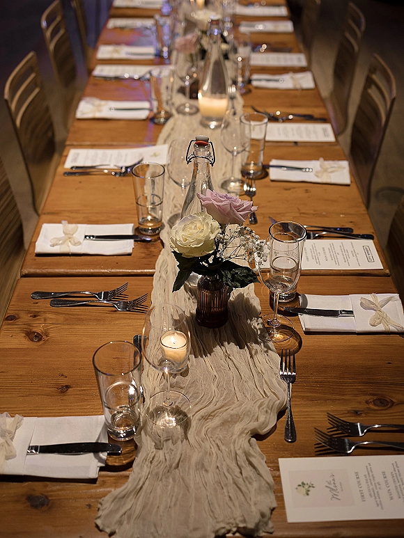 Reception tablescape on a farmhouse wedding table with linen runner, rose and baby’s breath bud vases, votive candles, and place cards in dim light