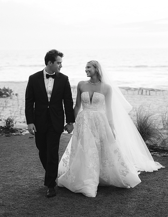 Couple portrait of bride and groom walking hand in hand on a beach, her long veil trailing behind near ocean dunes