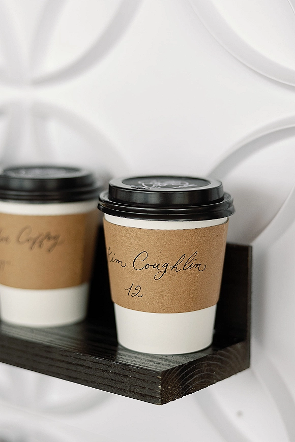Wedding coffee cups with custom coffee sleeves and handwritten calligraphy in a wooden holder against white wall tile background