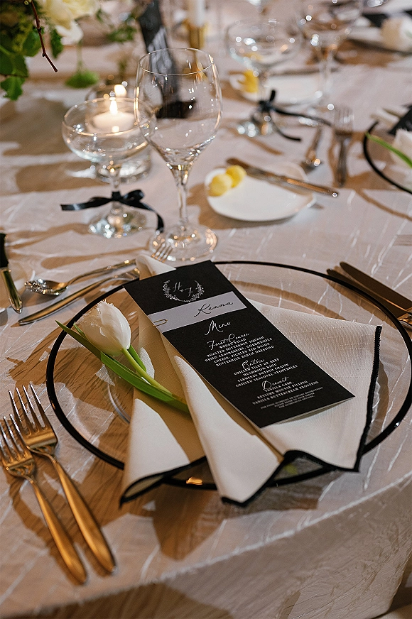 Reception tablescape with a black and white tablescape place setting, gold flatware, tulip, and floating candle amid softly blurred tables behind