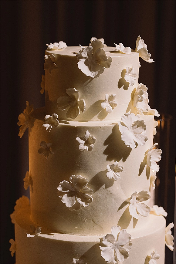 Wedding cake with white buttercream frosting, delicate sugar flowers, and subtle texture against a dark backdrop in warm lighting