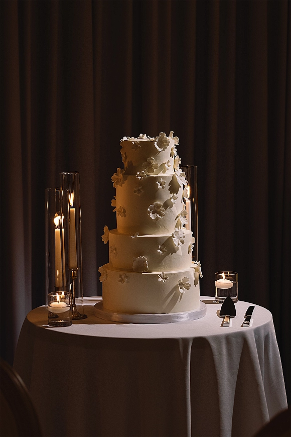 Wedding cake, tiered wedding cake in white with 3D sugar flowers on a stand, surrounded by taper and votive candles against dark drapes