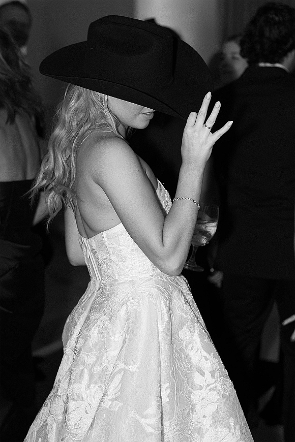 Bride portrait in cowboy hat, showing wedding ring in a strapless lace dress amid dimly lit indoor reception guests