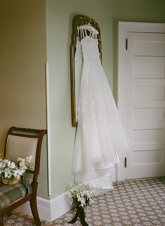 Wedding dress hanging from a mirror, strapless bridal gown with bouquet and ribbon on a chair in a simple bedroom setting