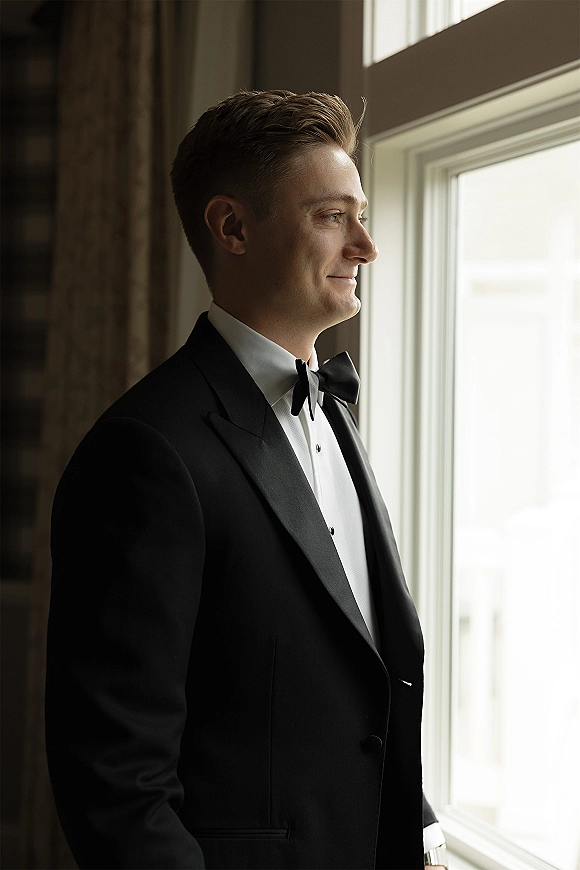Groom portrait in a black tuxedo, bow tie, and cufflinks standing by a window with curtains in a softly lit indoor room