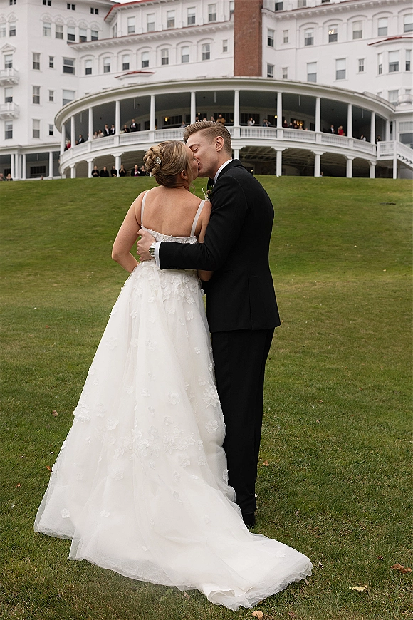 Wedding kiss portrait of bride and groom kissing on a lawn, her lace-back dress and updo visible, grand hotel veranda behind
