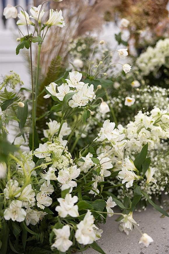 Wedding florals with white wedding flowers, baby's breath and fern fronds arranged along a stone floor in a softly blurred indoor setting