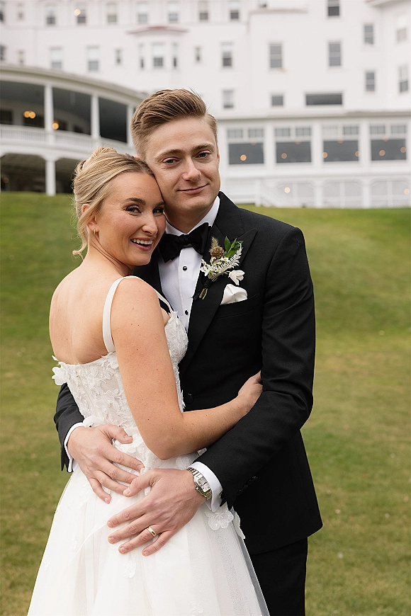 Couple portrait of bride and groom embrace, her lace bodice dress and his black tuxedo, on a lawn before a white building with balcony