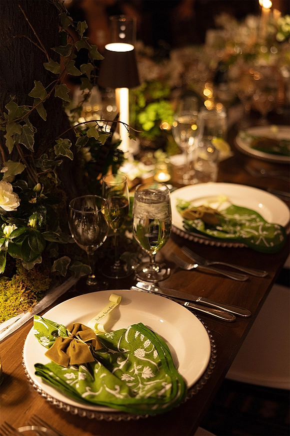 Reception tablescape with green wedding tablescape details, ivy garland, velvet napkin bows, candlelight and lamps on a wooden table under bistro lights