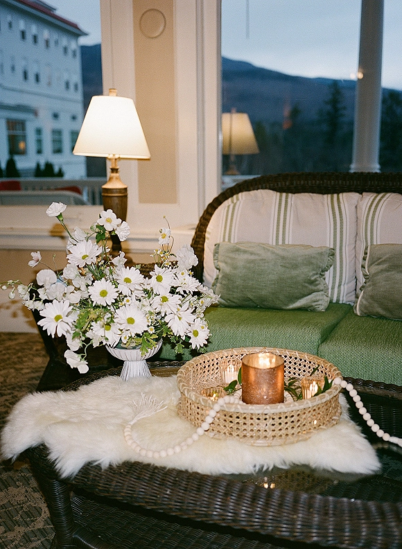 Wedding lounge seating with a wicker sofa and green cushions, candlelit coffee table and daisies on a covered porch with white columns at dusk