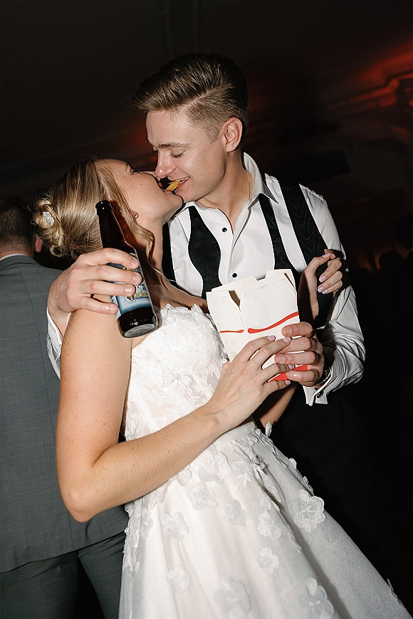 Wedding reception moment of bride and groom candid sharing fries from takeout boxes, bride holding a beer in a dim room with guests