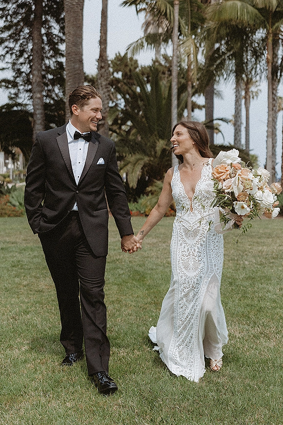 Couple portrait of bride and groom walking hand in hand, bride holding bouquet in lace dress beside palm trees and tropical lawn