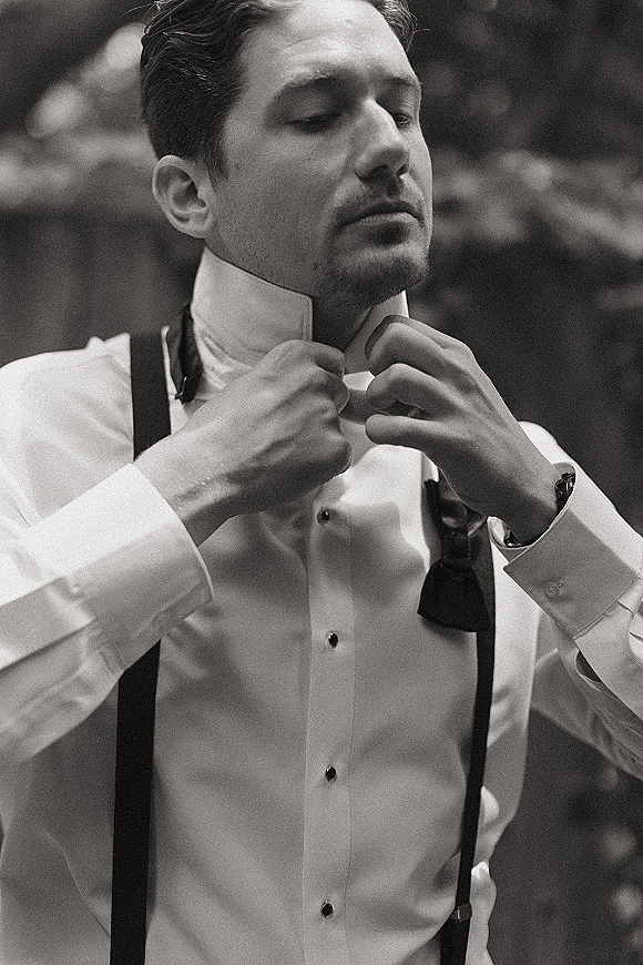 Groom getting ready, adjusting a black bow tie over a white dress shirt with suspenders, cufflinks and wristwatch, outdoors by blurred greenery