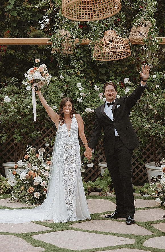Recessional moment as bride and groom walking hand in hand, bride raising bouquet, groom waving under hanging wicker lanterns in a vine garden aisle