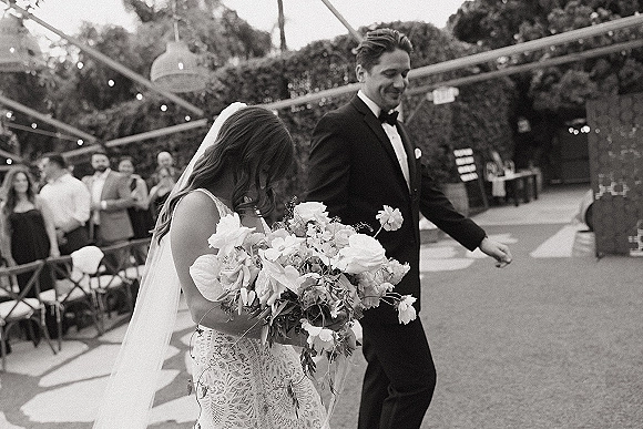 Wedding recessional as bride and groom walk the aisle, bride holding bouquet and long veil, guests seated under string lights outdoors