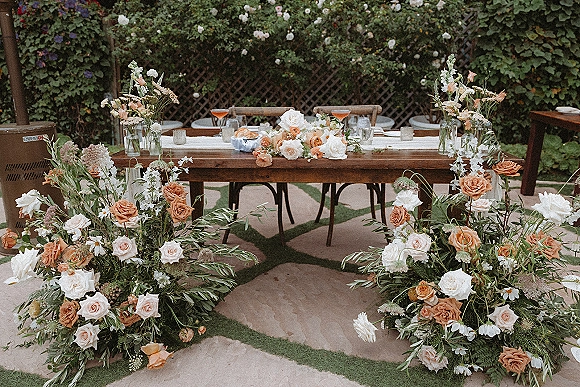 Reception sweetheart table with sweetheart table flowers of roses and greenery garland, votive candles and glasses on a patio by a flowering hedge