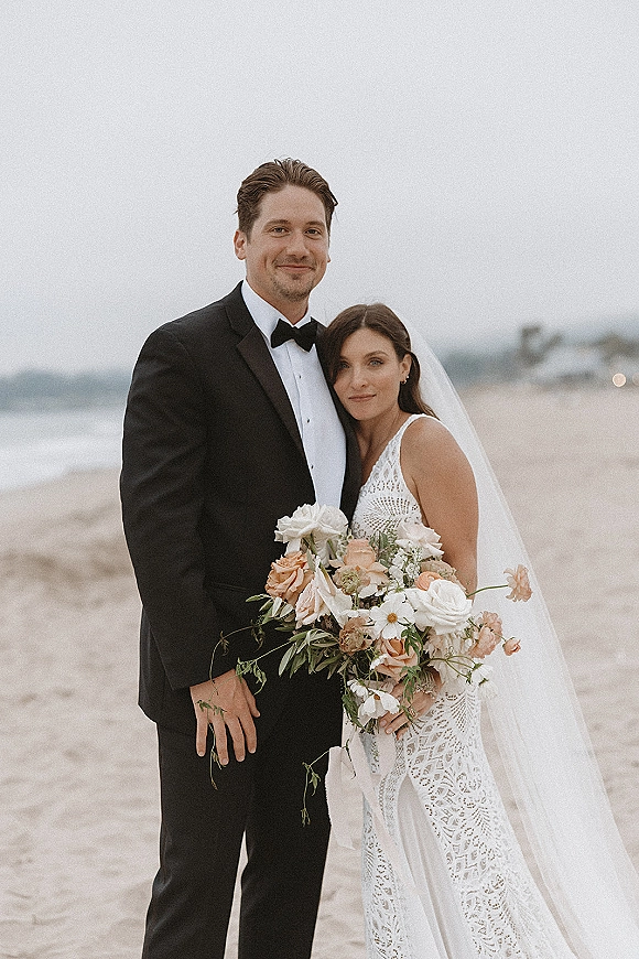 Couple portrait of bride and groom in a beach wedding portrait, she holds a bouquet and veil by the ocean under overcast sky