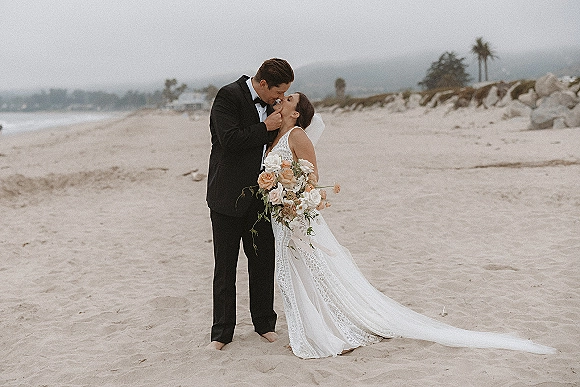 Wedding couple portrait of bride and groom kissing on a sandy beach, her lace dress and veil flowing, holding rose bouquet by shoreline