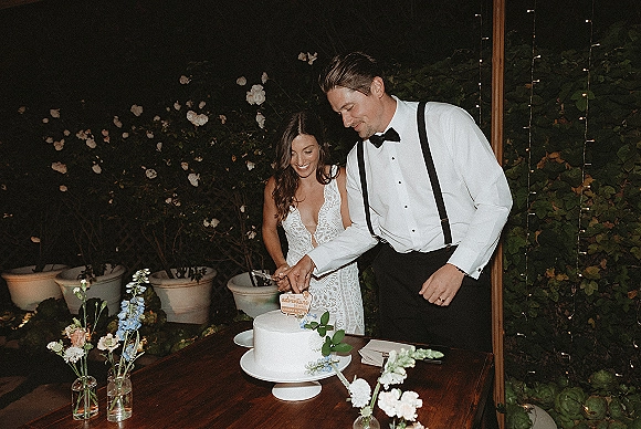 Wedding cake cutting as bride in lace dress and groom in suspenders slice a single-tier cake under string lights in a night garden