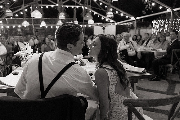 First dance moment as bride in a lace wedding dress slow dances with groom in suspenders under bistro string lights in a tent at night