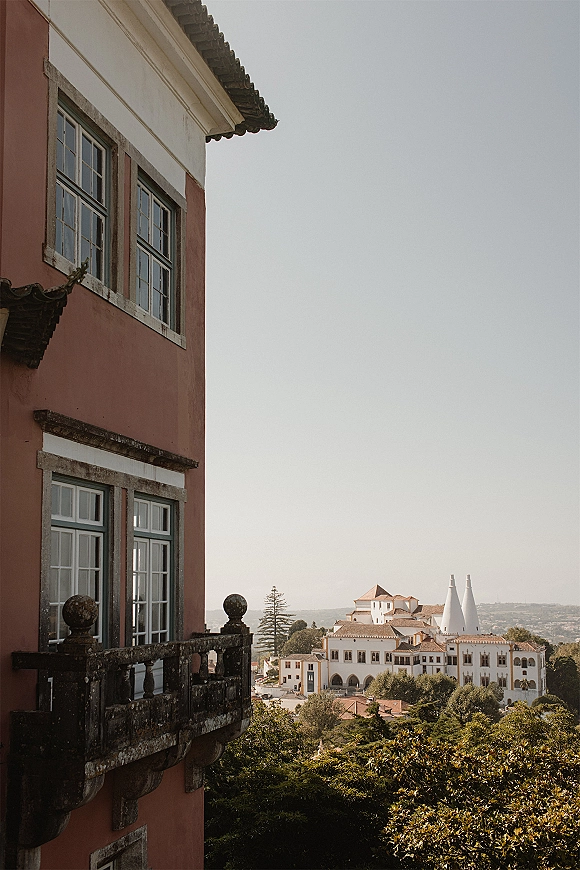 Wedding venue exterior with a balcony and stone railing, grand palace facade rising above treetops on a hillside under open sky