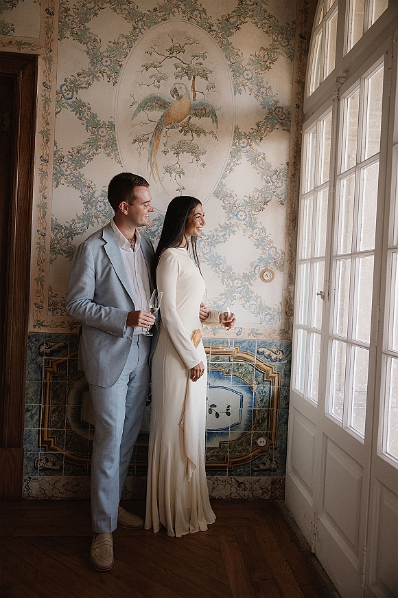 Couple portrait of bride and groom by window, holding champagne flutes in soft natural light against a painted mural wall and wallpaper